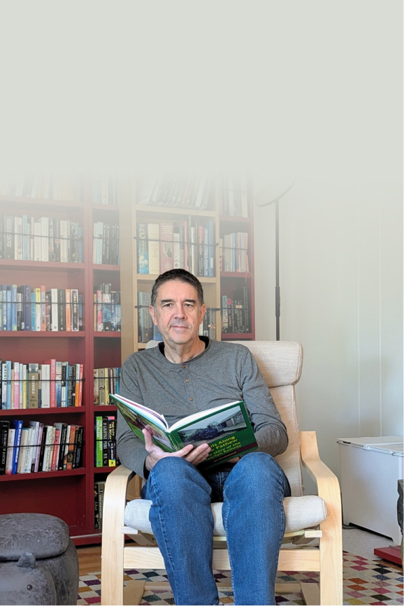 Dan Quine sits in a wooden armchair reading a book in a brightly lit room filled with red bookshelves packed with books.