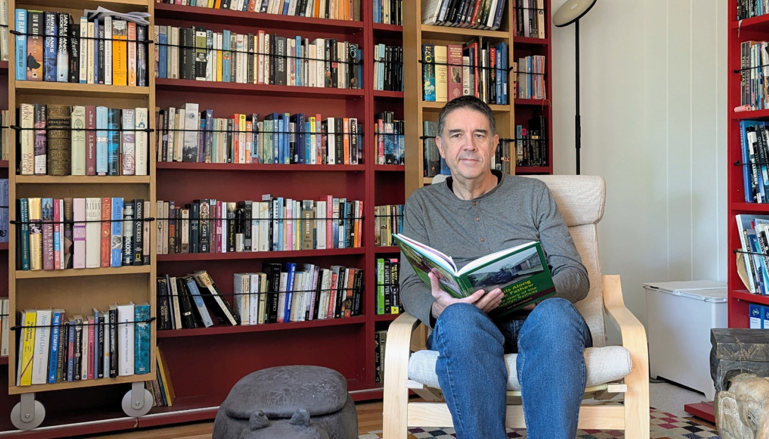 Dan Quine sits in a wooden armchair reading a book in a brightly lit room filled with red bookshelves packed with books.