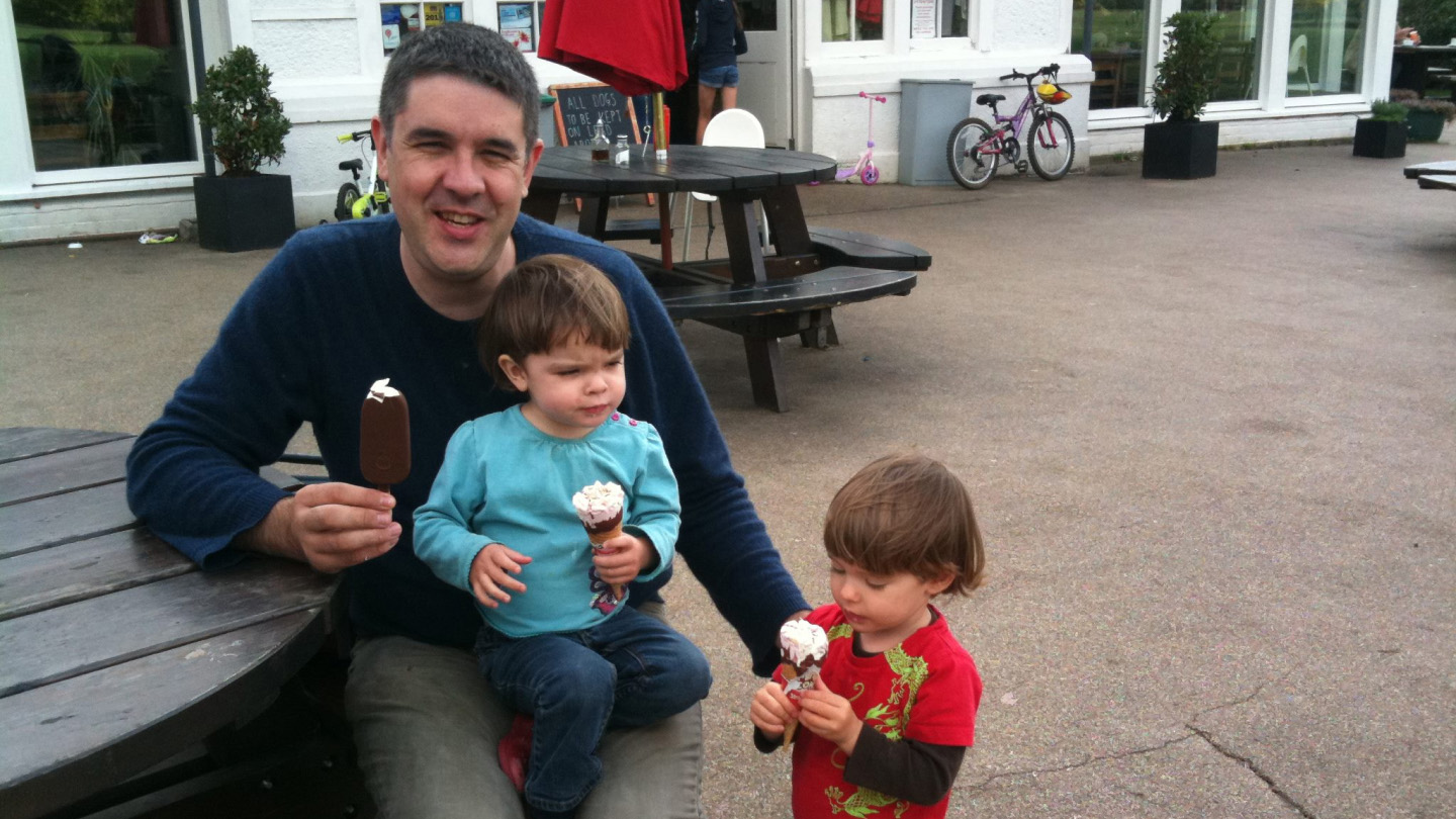 Dan Quine sits at an outdoor picnic table holding a chocolate-covered ice cream bar, with a young child in a blue shirt and pink shoes holding a small ice cream cone, while another child in a red shirt stands nearby eating their own ice cream, with a background of a café and bicycles.