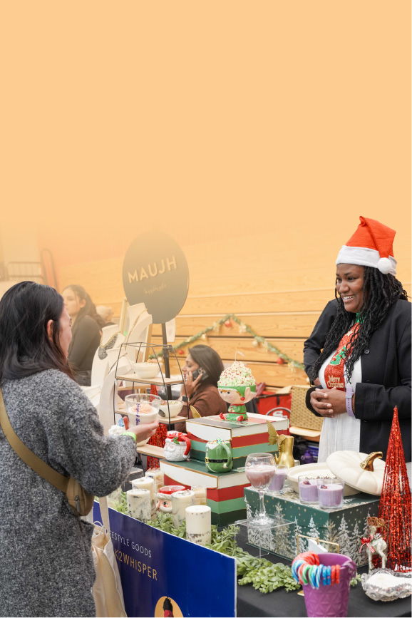 A business owner in holiday attire stands behind a table display of her handmade candles as she smiles at a shopper.