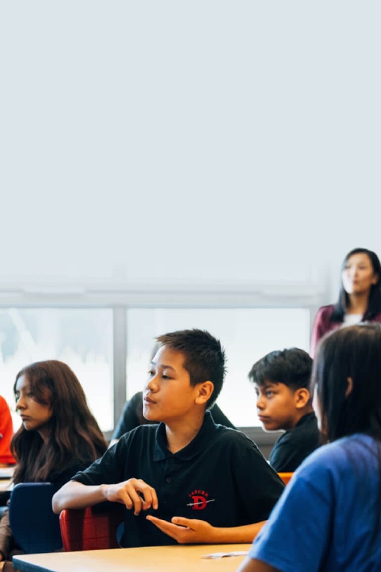 Middle school students engaged in classroom discussion, with a teacher observing in the background.