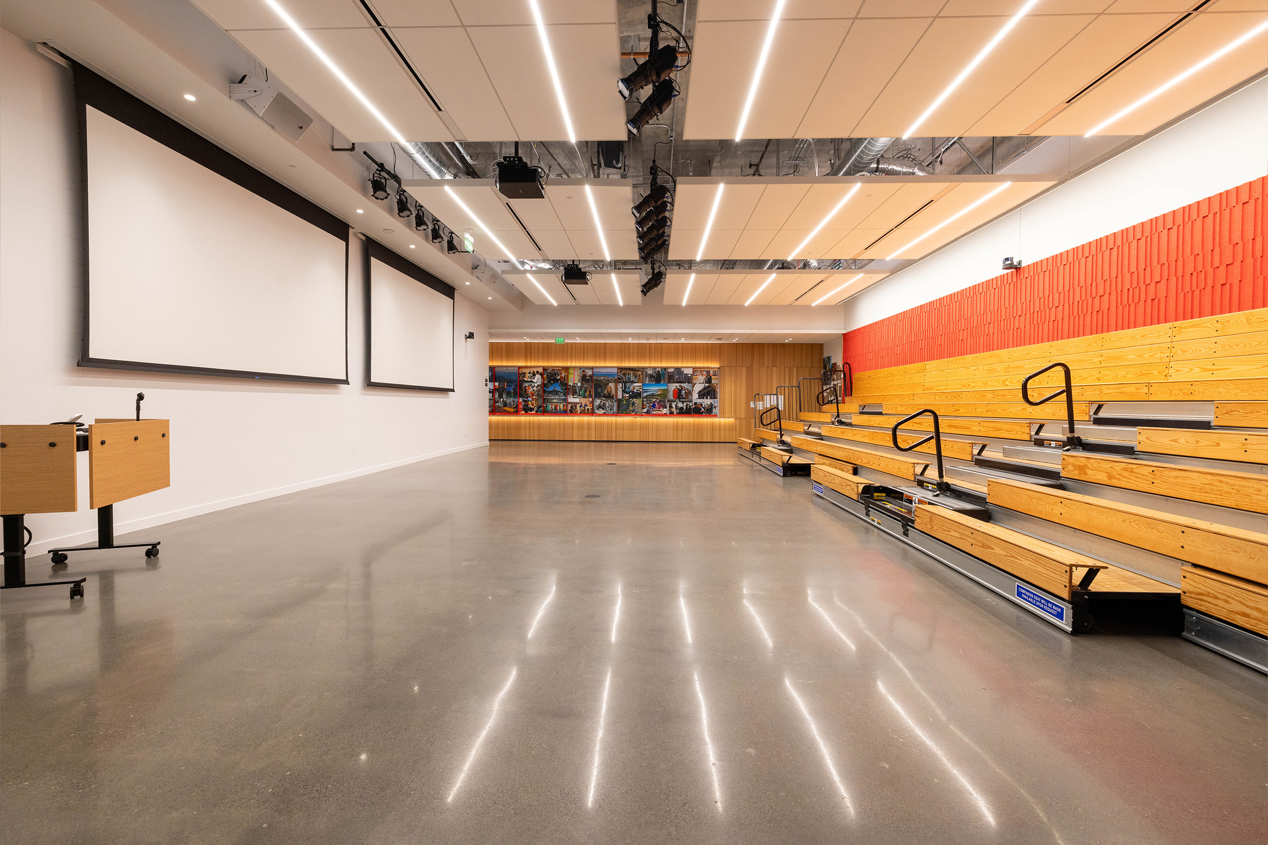 Large empty room with projector screens, podium and stadium seating on one wall.
