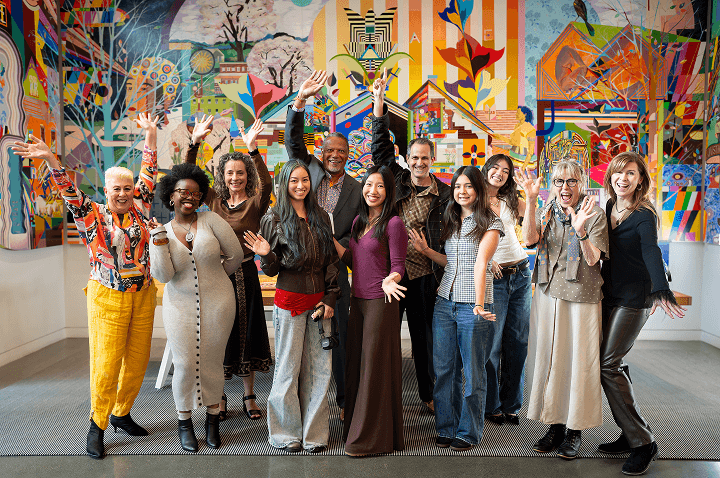 A group of people stand in front of a colorful mural in the CZI Community Space, smiling and raising their hands in a celebratory pose.