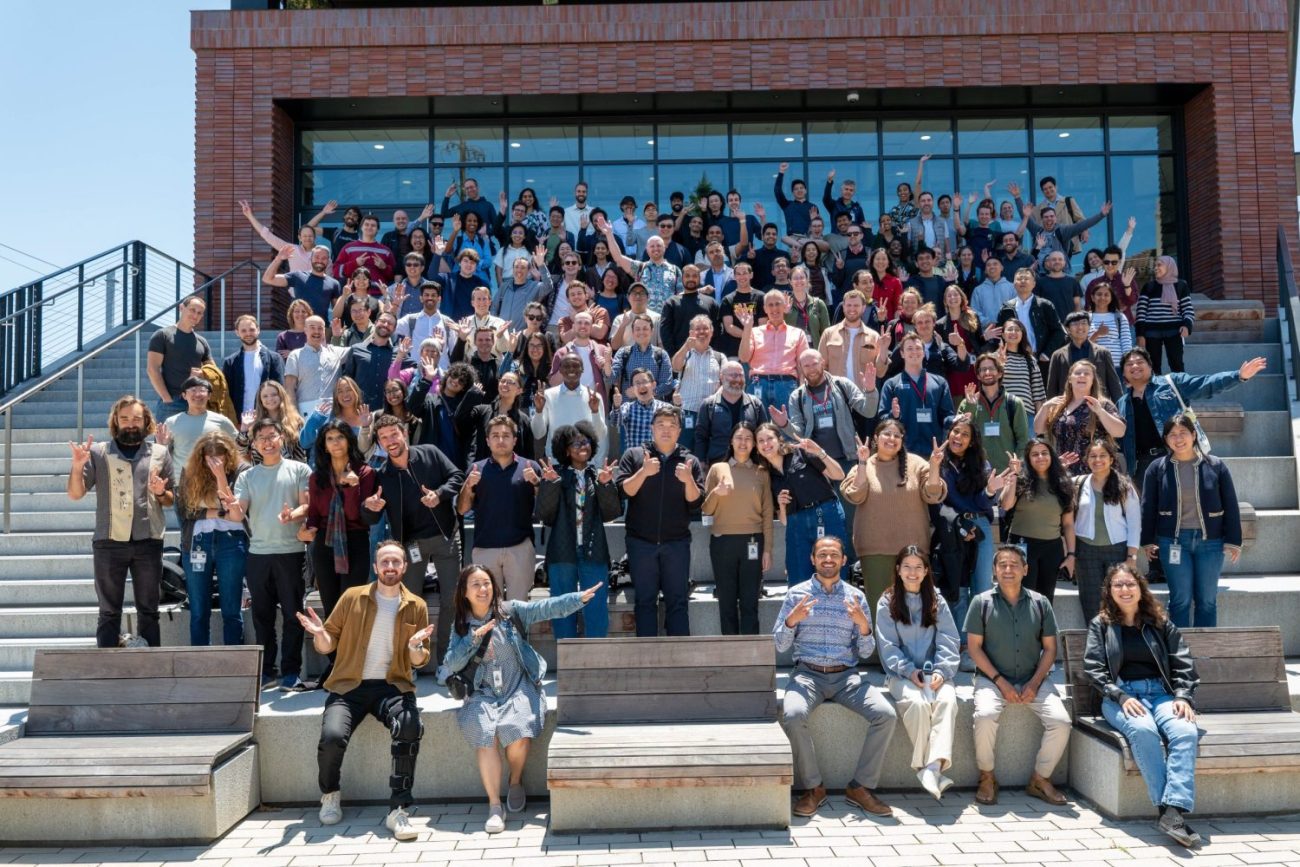 Large group photo of CZI staff gathered outdoors on tiered seating in front of the Redwood City headquarters, smiling and waving toward the camera on a sunny day