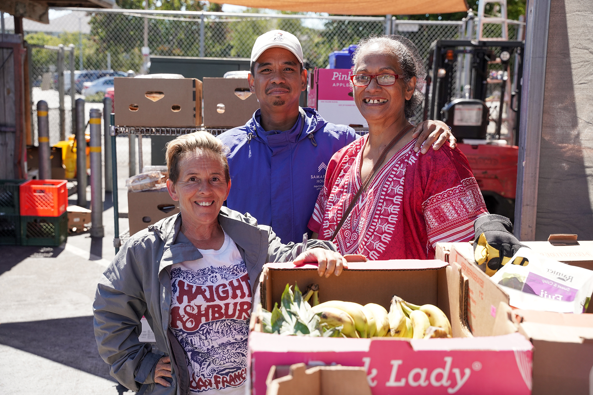 Three people standing together smiling in front of vegetable and fruit boxes used to prepare meals for their community.