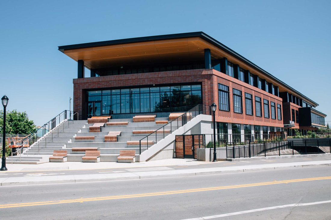Exterior view of a modern brick and glass building with tiered concrete seating and wooden benches, located across a street under a clear blue sky.