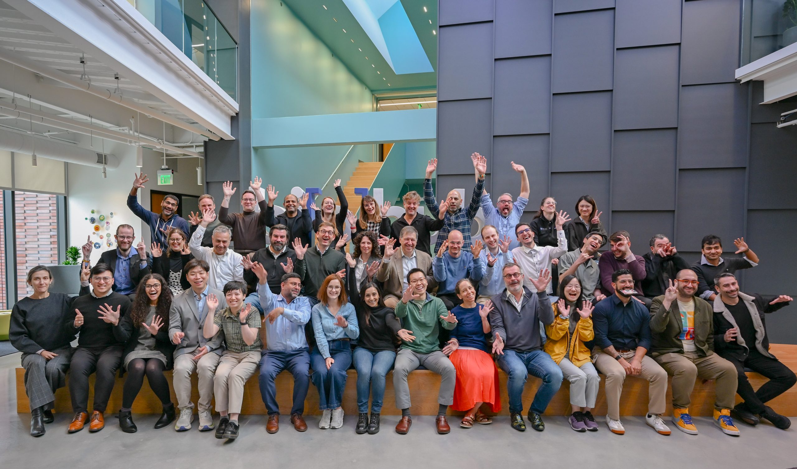 Participants of the Microglia Taxonomy Working Group Meeting waving and cheering in front of the Bi[o]hub sign in the communicating space of the CZI headquarters in Redwood City, CA.