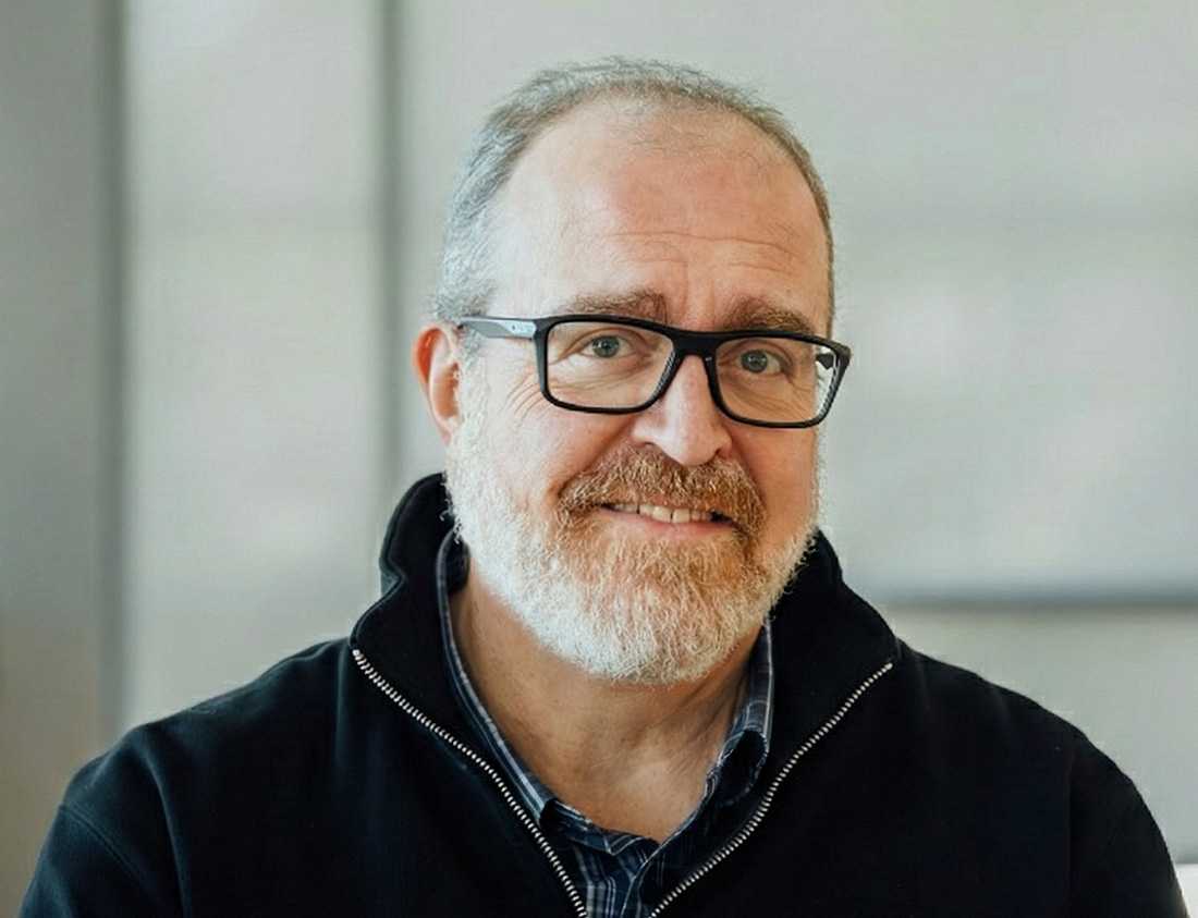 Portrait of Marc Malandro smiling, wearing glasses and a dark zip-up sweater, seated in a bright, modern office setting.