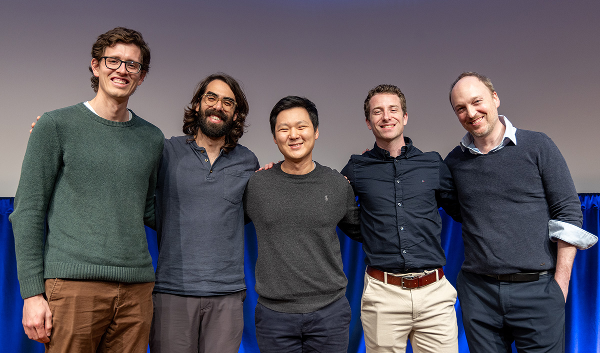 Five speakers standing together on stage at a symposium, smiling with arms around each other in front of a blue curtain.