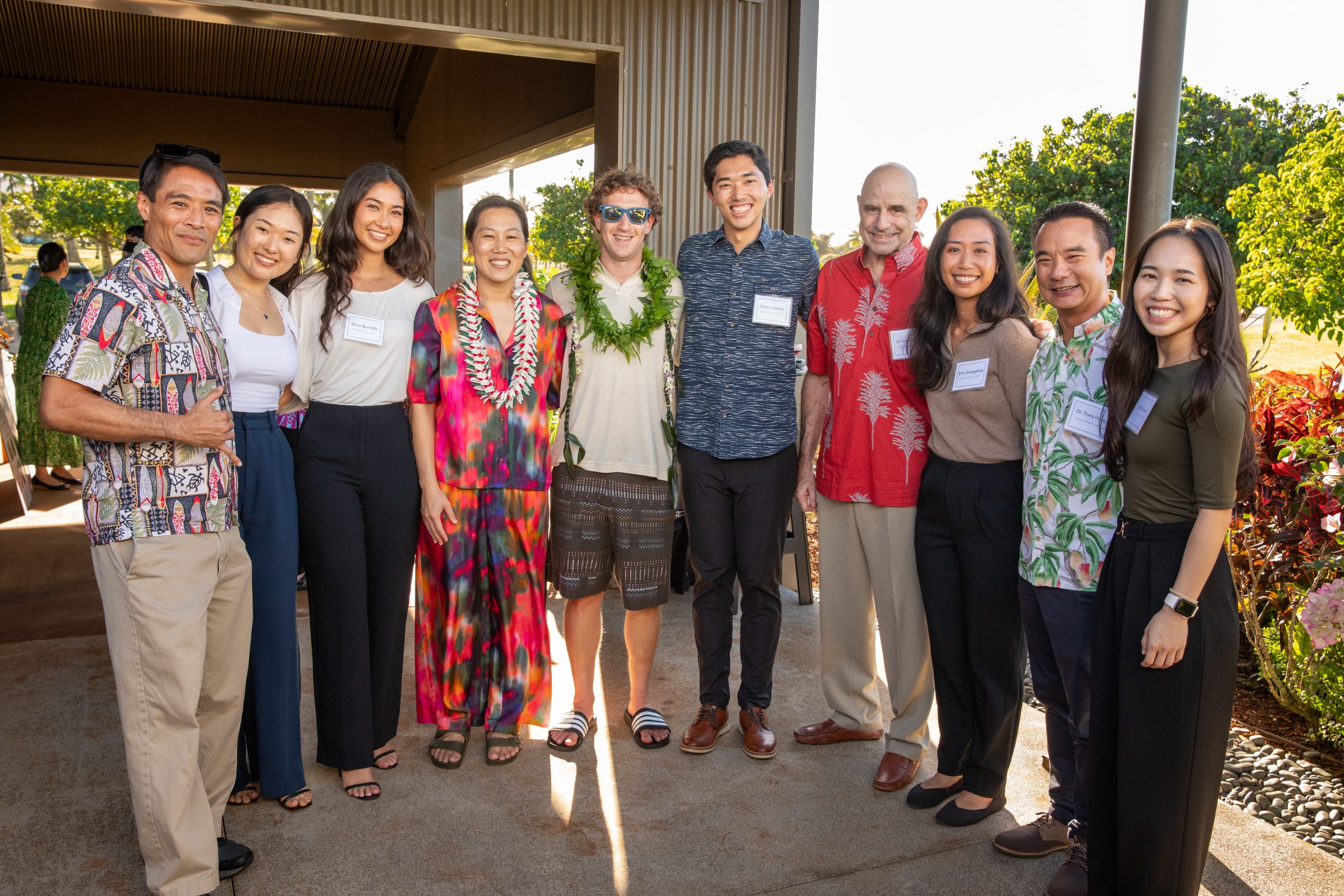 A group of nine smiling people posing together outdoors at a casual event, some wearing colorful Hawaiian attire and leis.