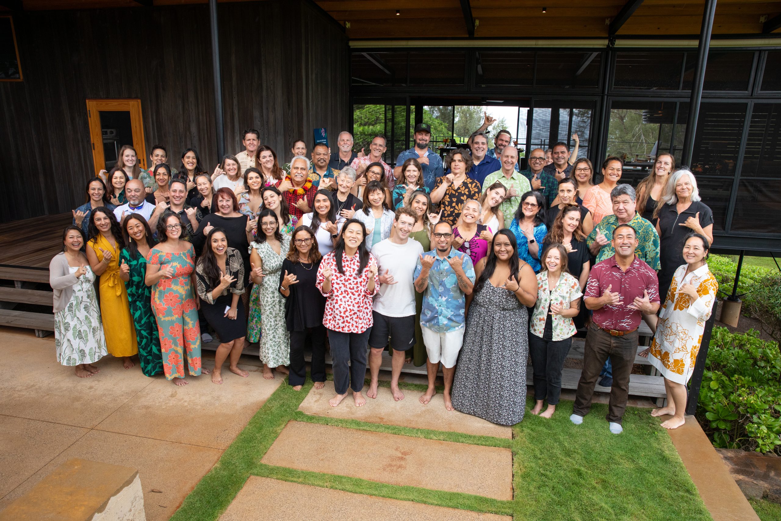 A large group of approximately fifty people smiling and posing together outdoors in front of a wooden building.