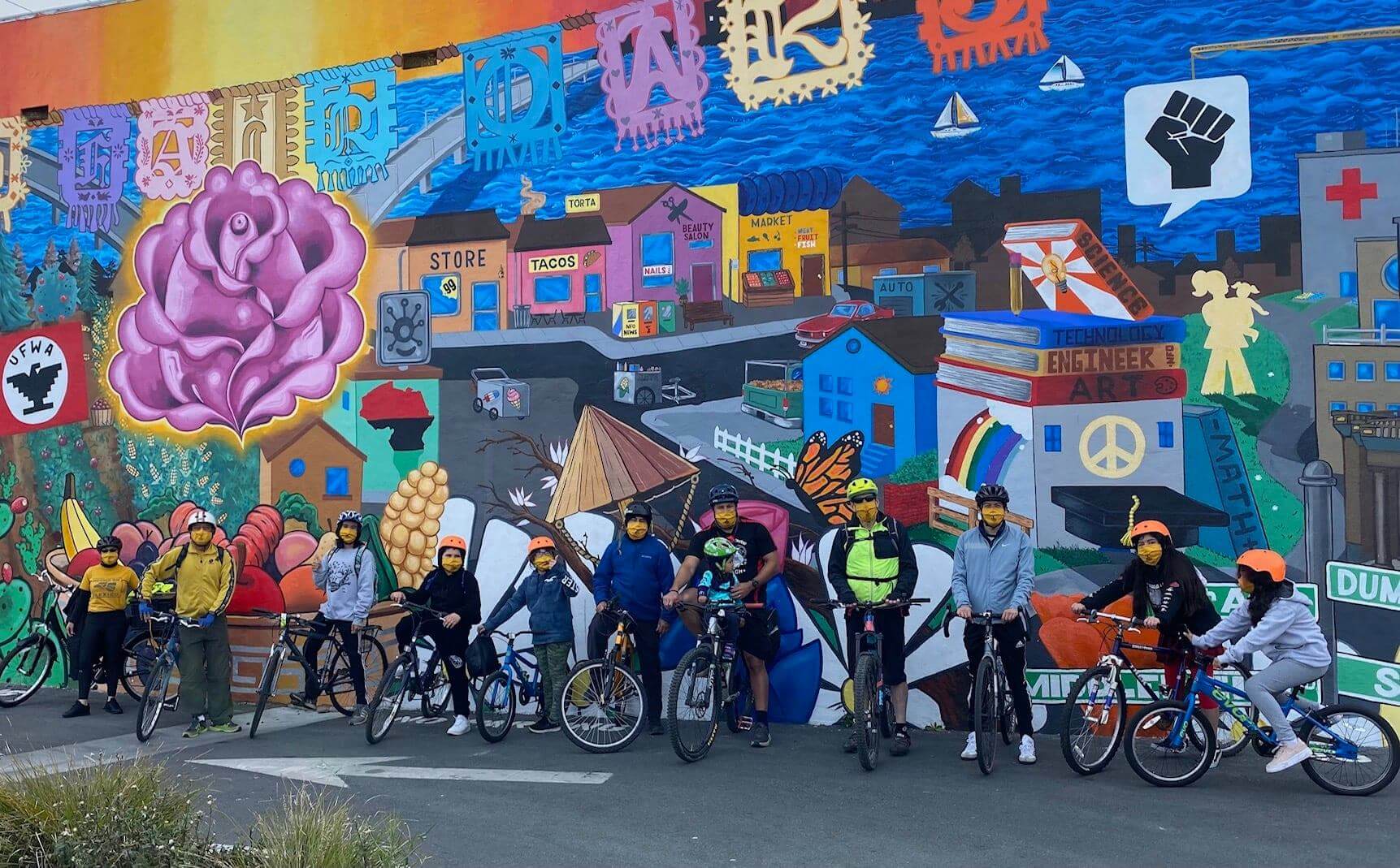 A group of community member sit on their bikes in front of a colorful mural.