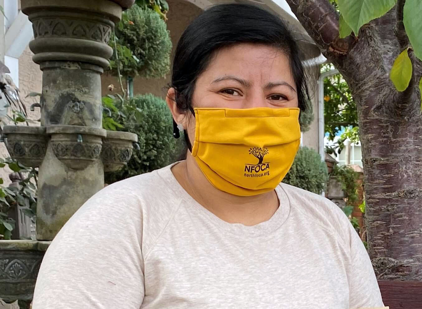 A woman in a tan shirt wears a yellow face mask and holds a brown bag with the NFOCA logo.