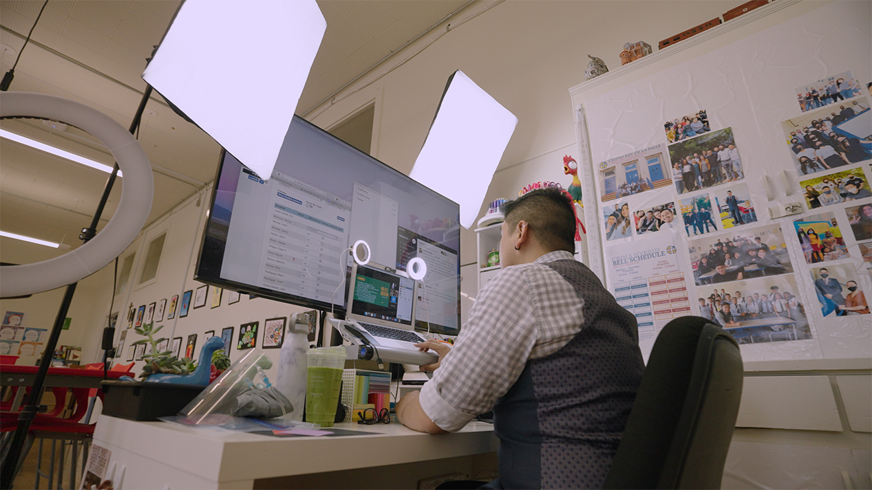 A teacher sits at his desk with a laptop and large screen in front of him for virtual instruction of his class.
