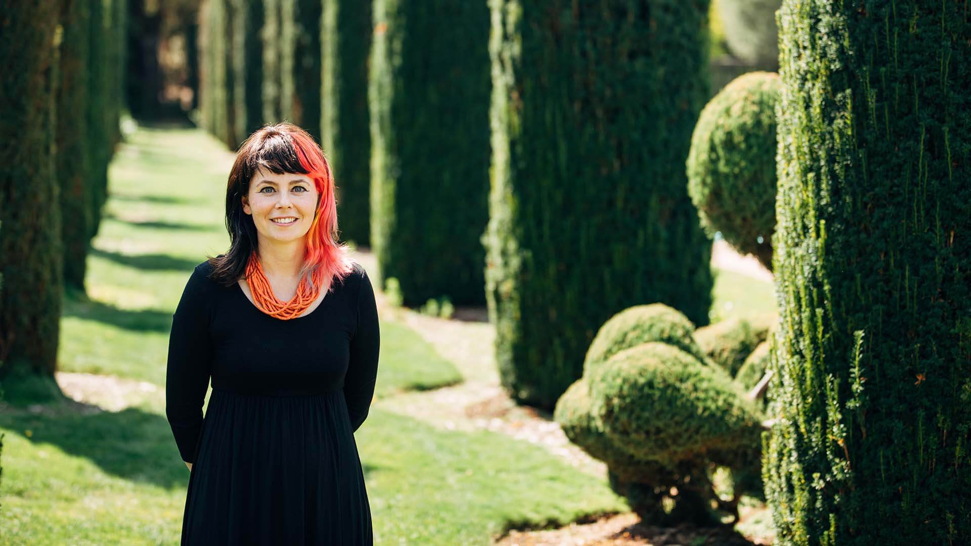 A women with pink and black hair smiles with her hands behind her back. Columns of greenery are behind her.