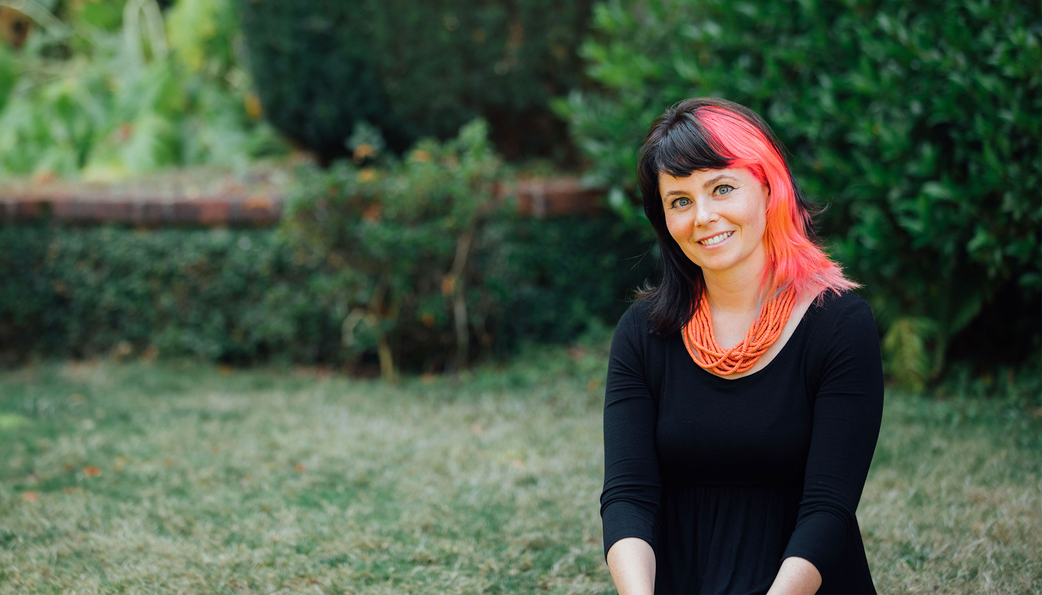 A women with black and pink hair smiles with greenery behind her.