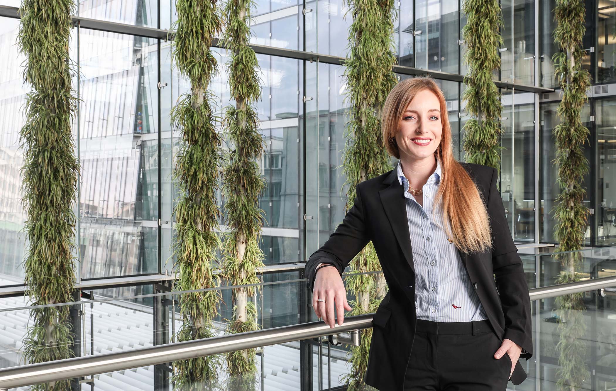 A women is dressed in business attire and leans again a railing in a glass building.