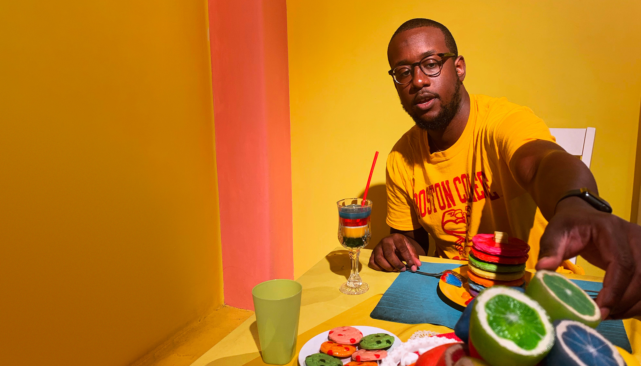 A man wearing a yellow shirt reaches for a bright green lime on a table with other colorful treats.