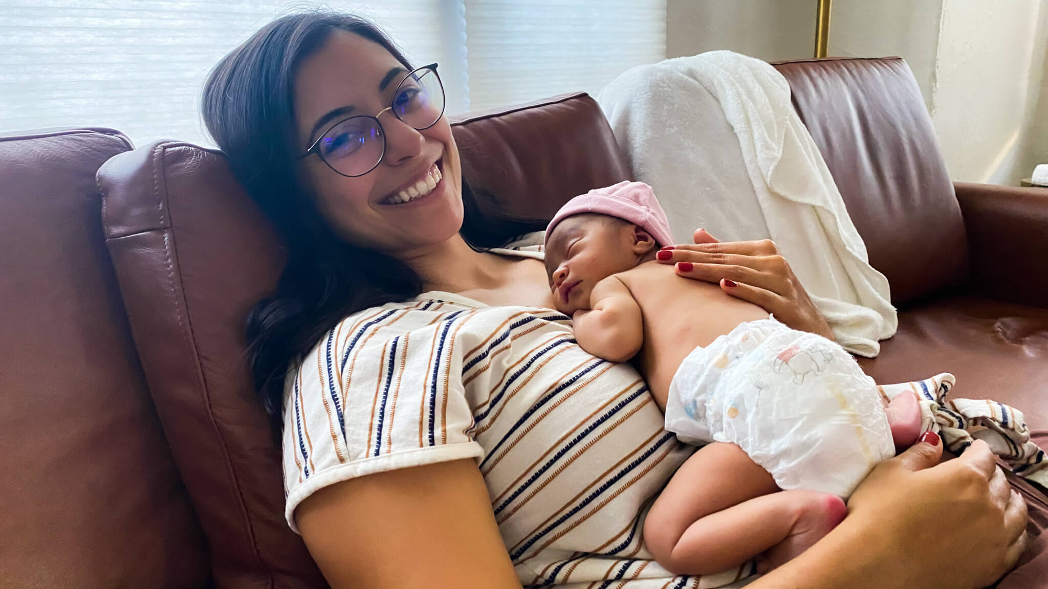 A woman lays on a couch smiling and holds a sleeping baby.