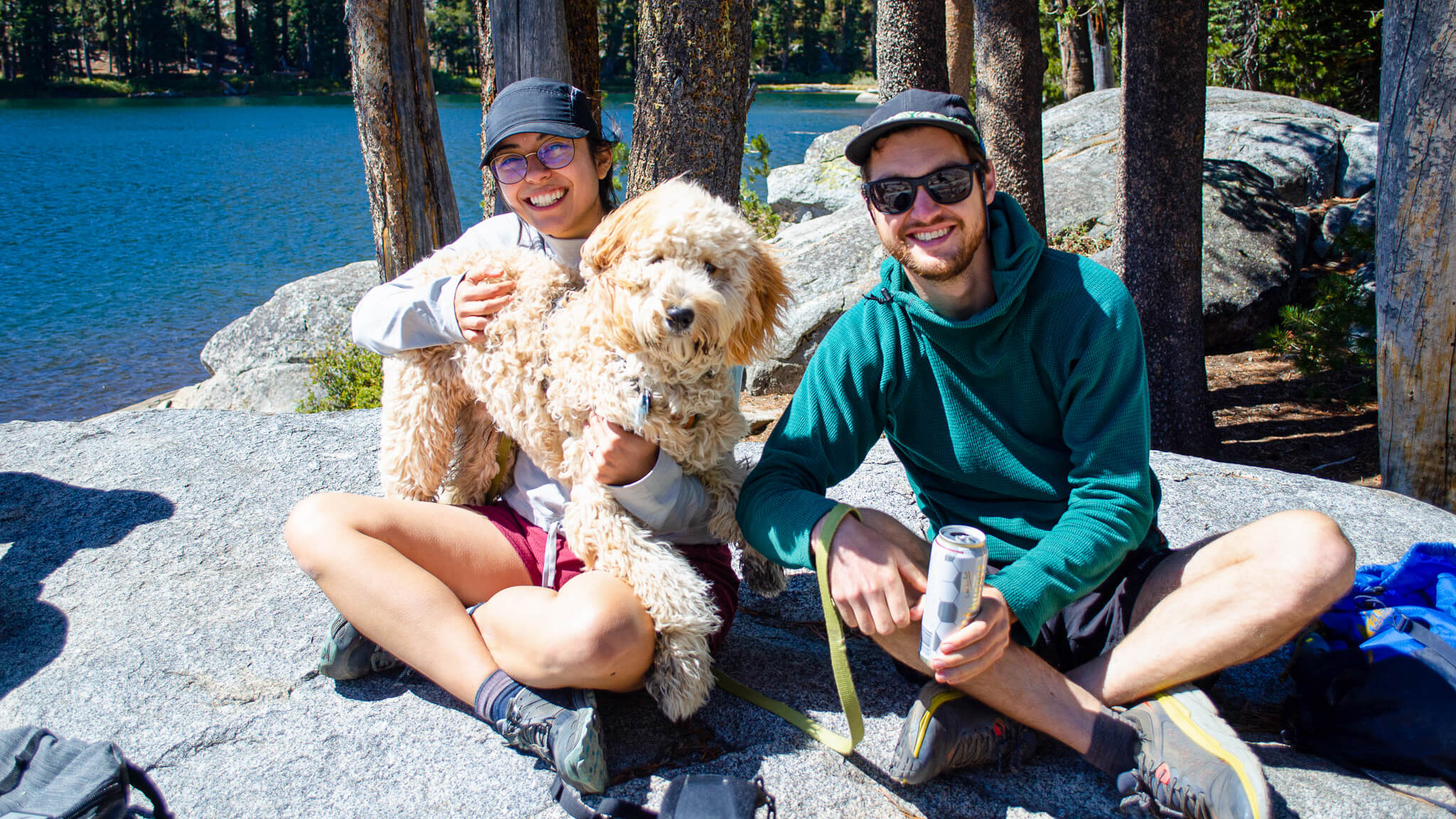 A woman and man sit on a rock holding a white, fluffy dog. Water and trees surround them.