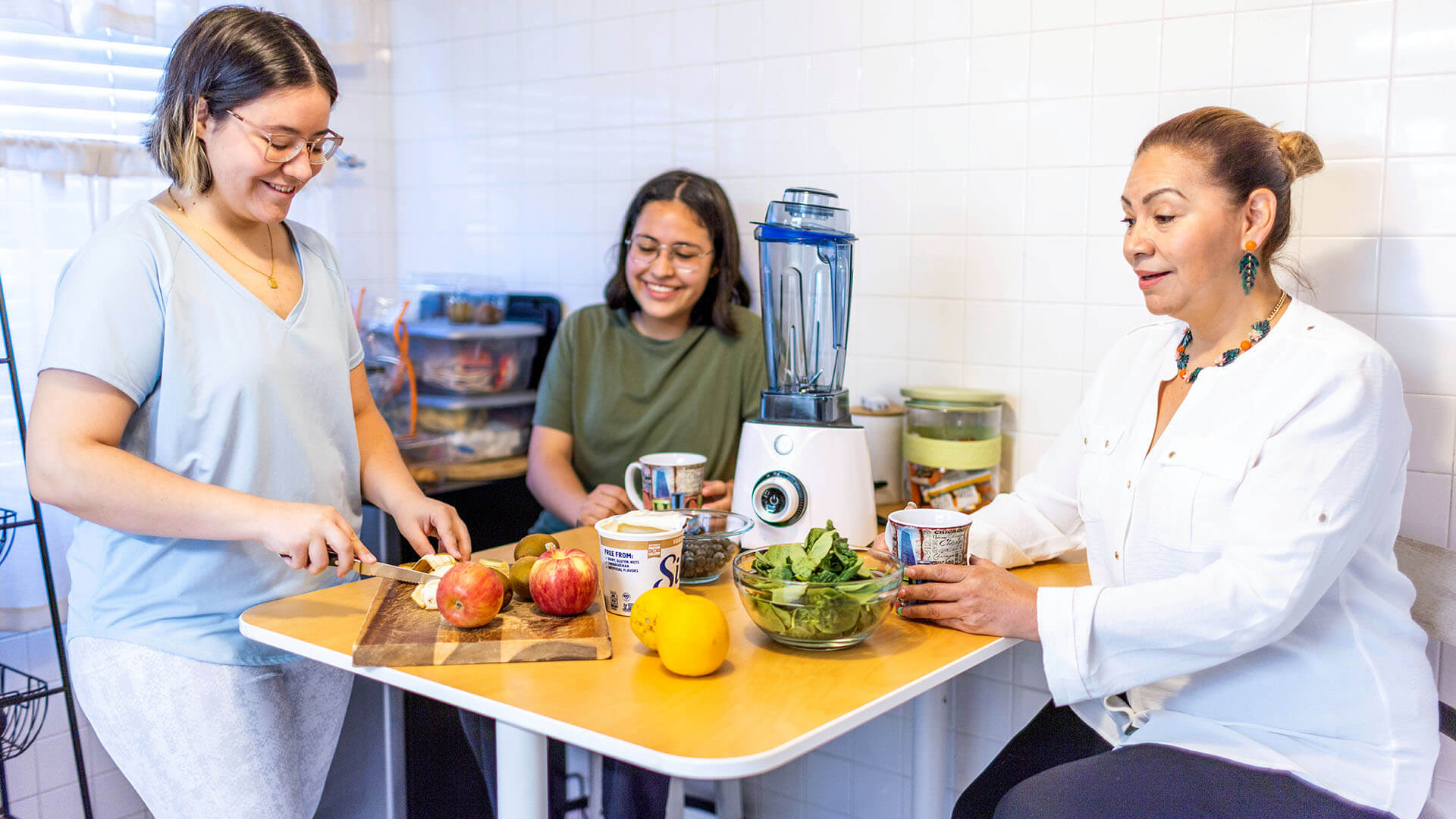 Three women talk and smile in the kitchen. One is cutting fruit for a smoothie while the others watch.