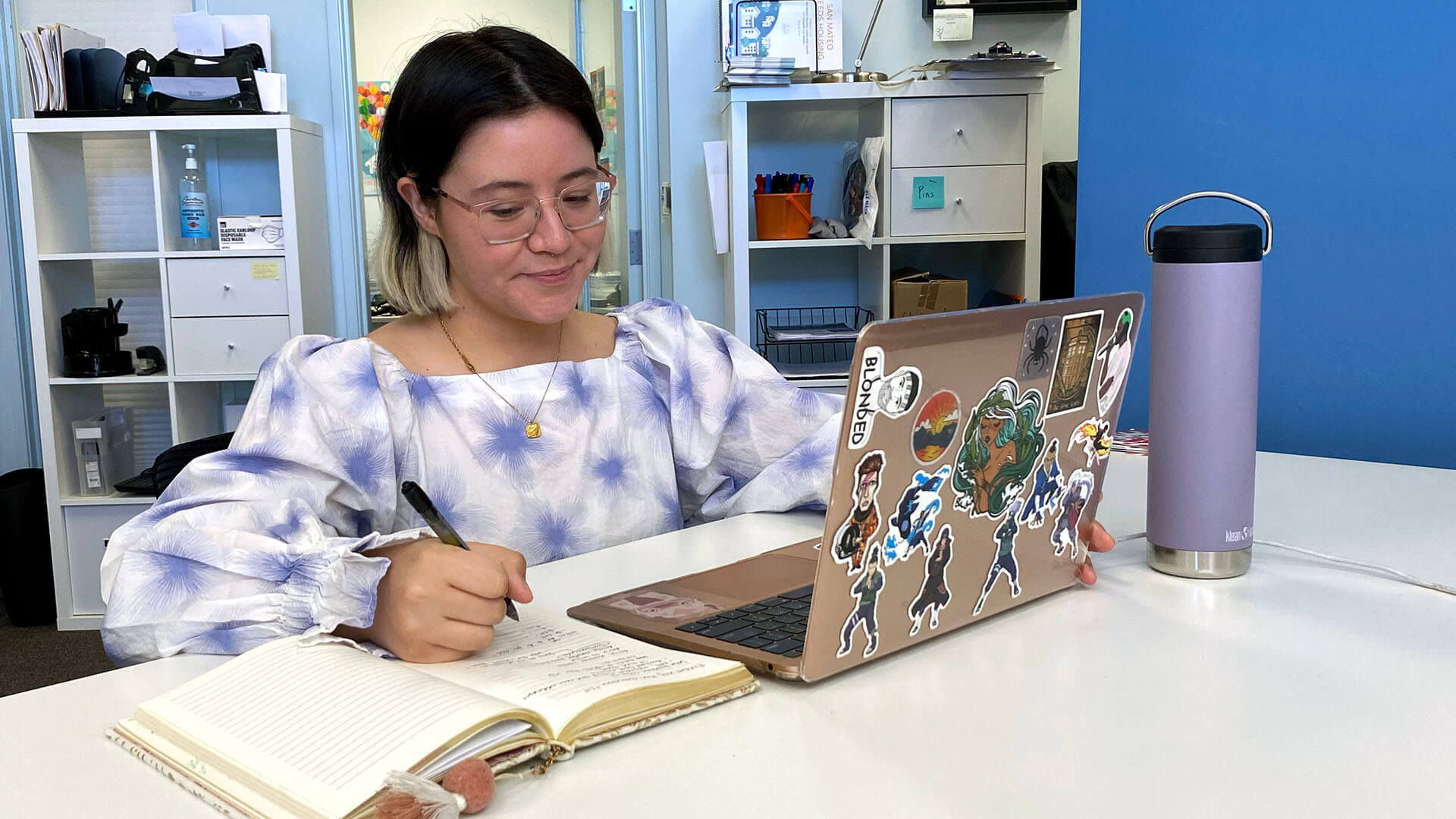 A woman wears a puffy, purple and white blouse and sits at a desk writing in a notebook.