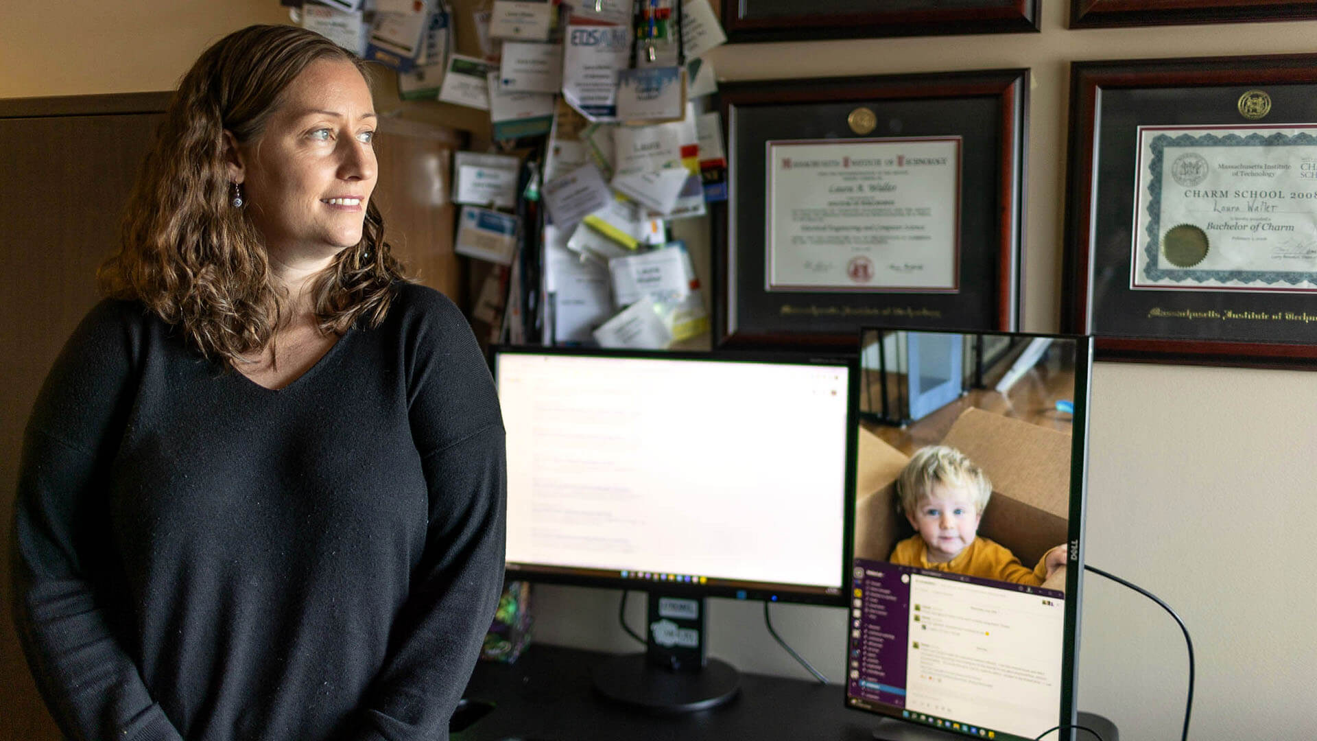 Laura sits on the edge of a desk. On top of the desk are three computer screens.