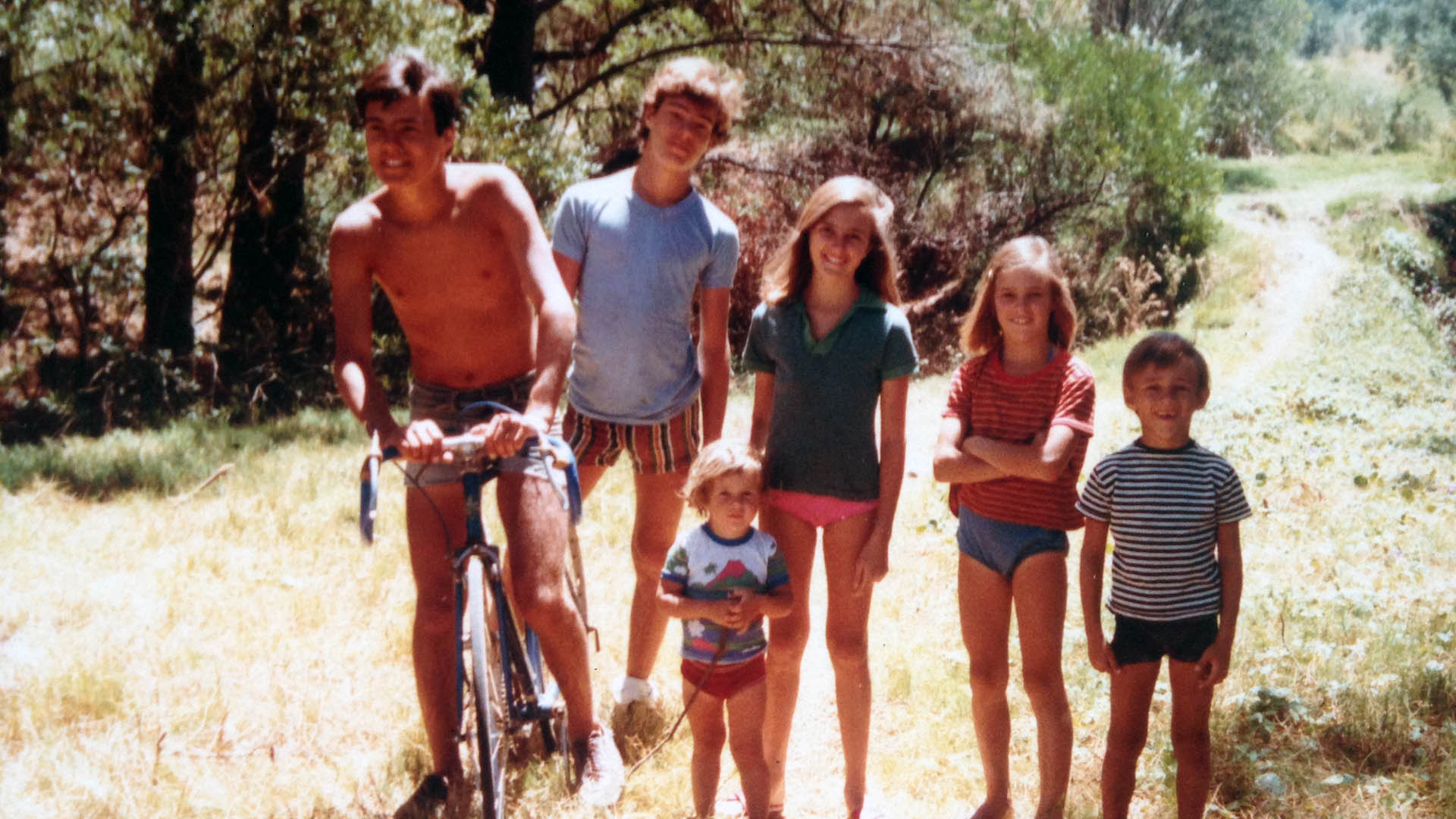 A group of young siblings smile for a photo outside. One sits on a bike. 