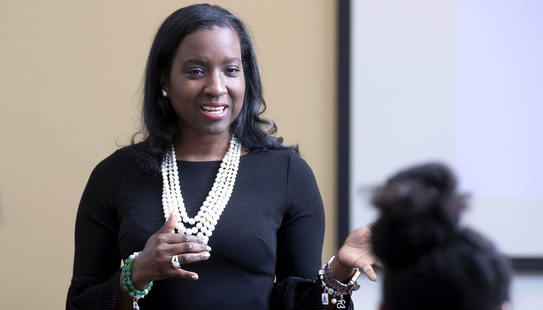 Andrea Lewis wears white pearls and a black dress while speaking in front of an audience. 