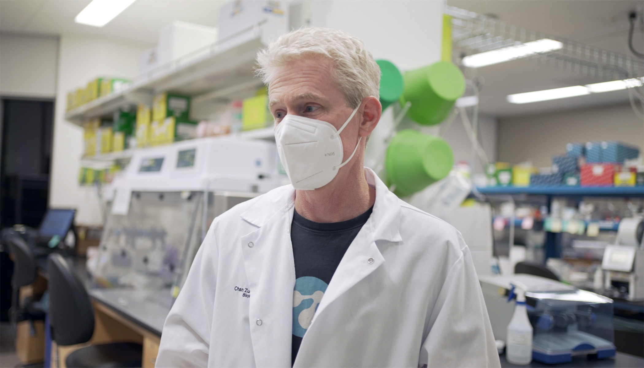 Man stands in lab. He is wearing a white mask and a white lab coat. 