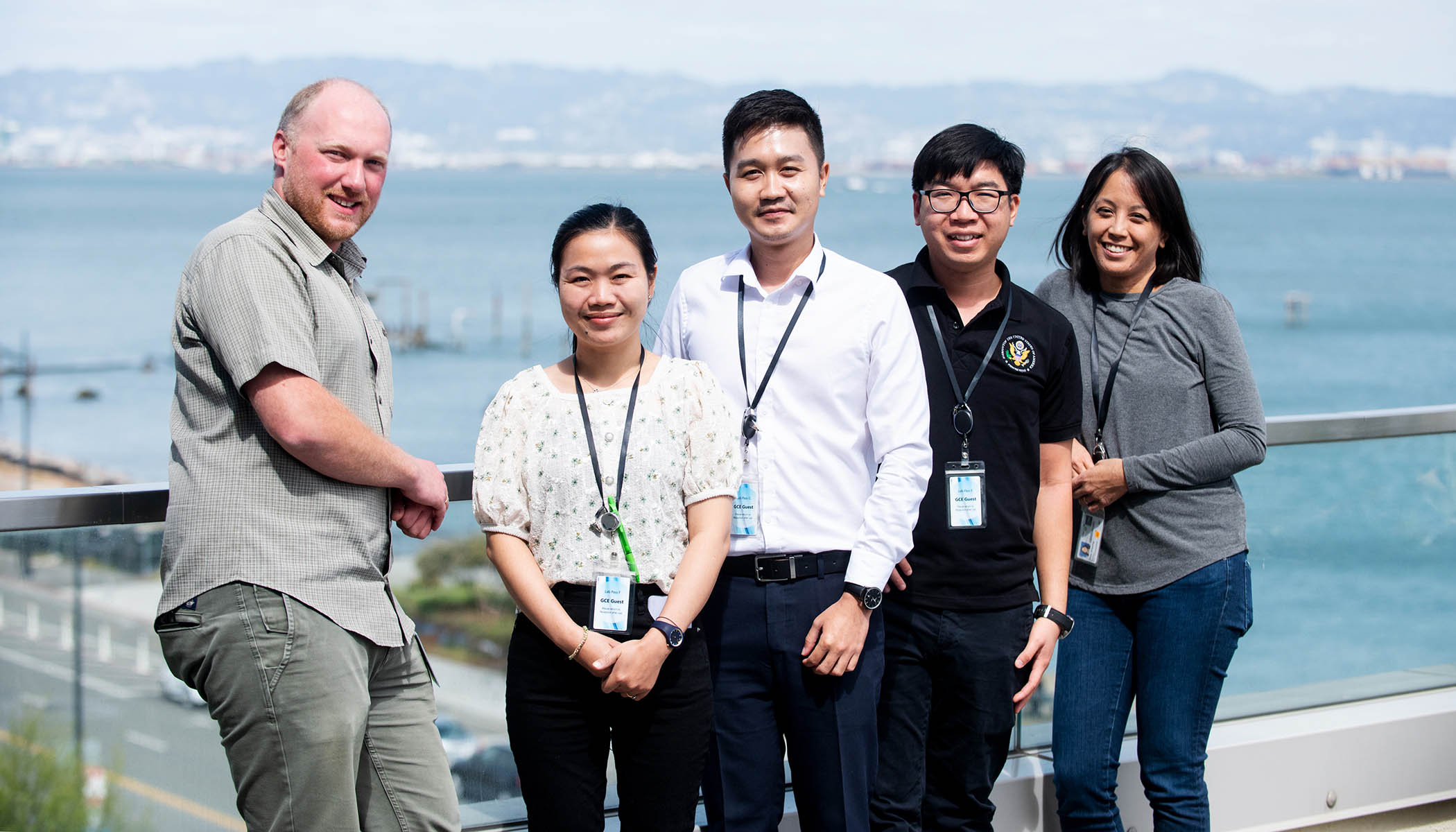 A group of five individuals stands together, smiling. They are leaning against a glass barrier, in the background is the ocean.
