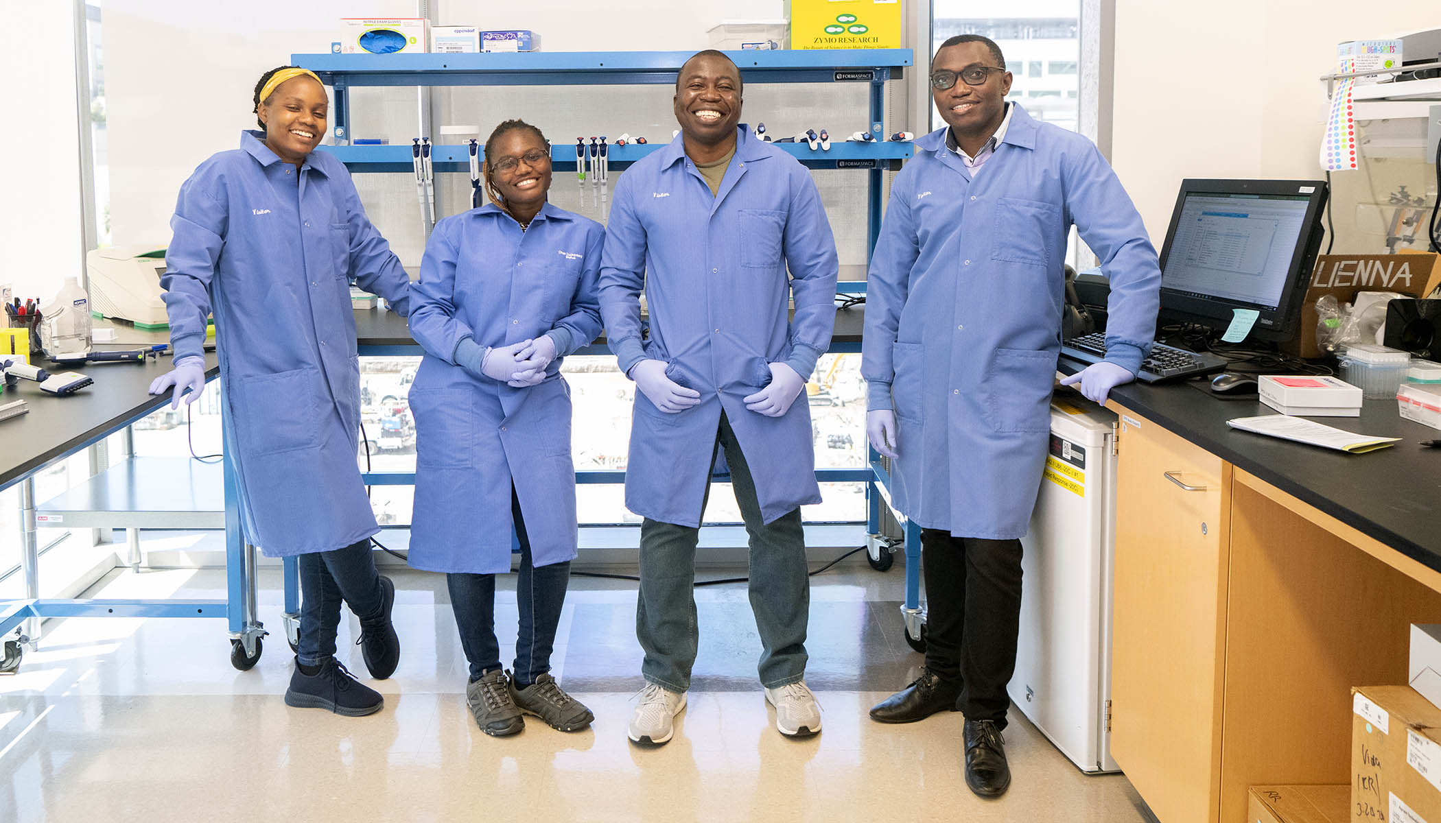 Four individuals stand together in a lab, all wearing blue lab coats. 