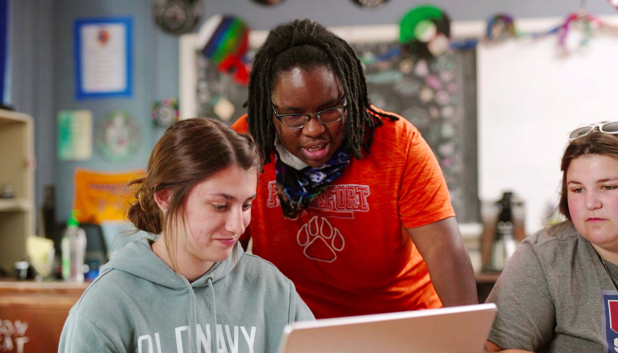 A teacher and two students look at a laptop in a classroom.