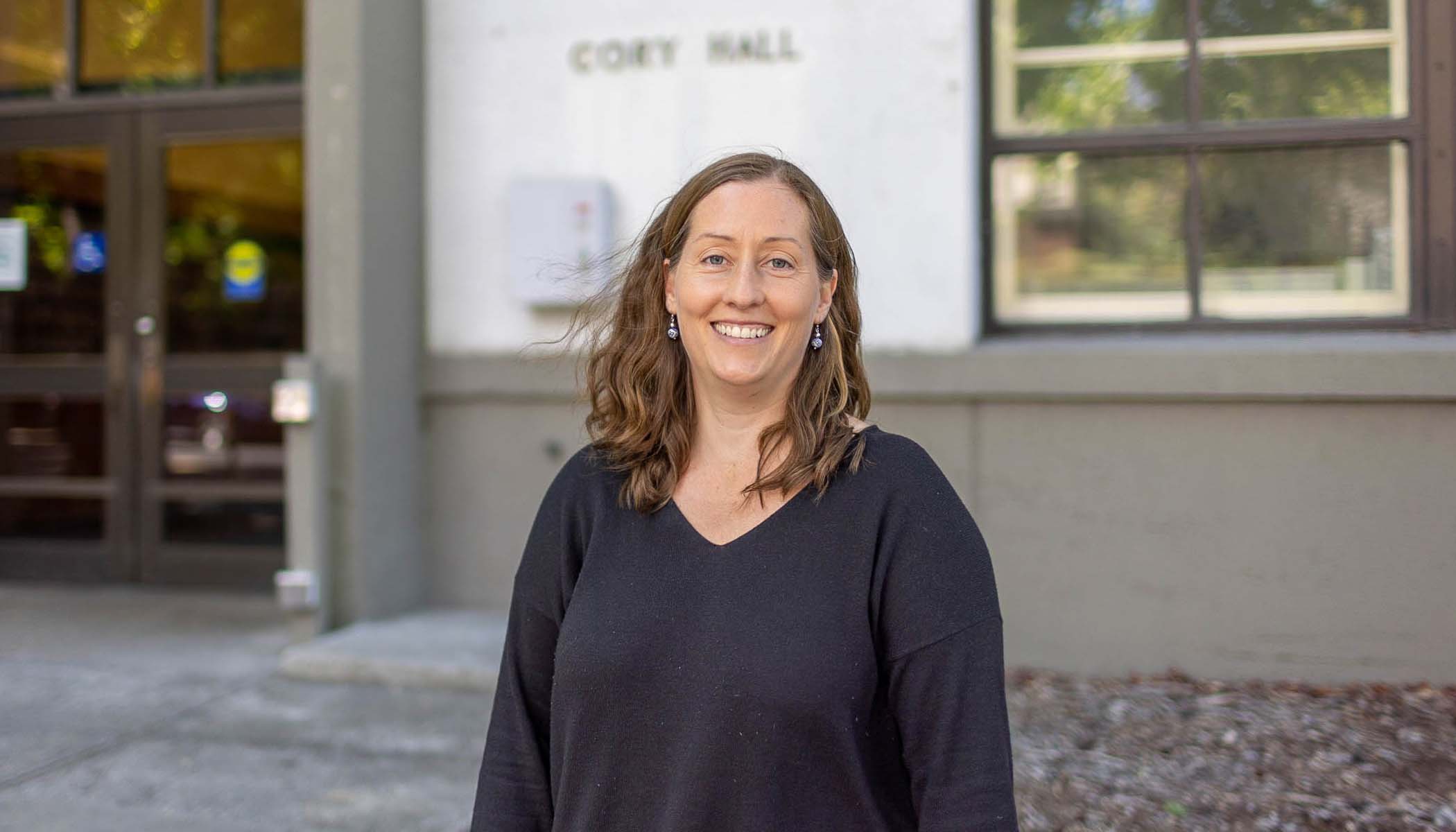 A woman in a black shirt smiles in front of a college building.
