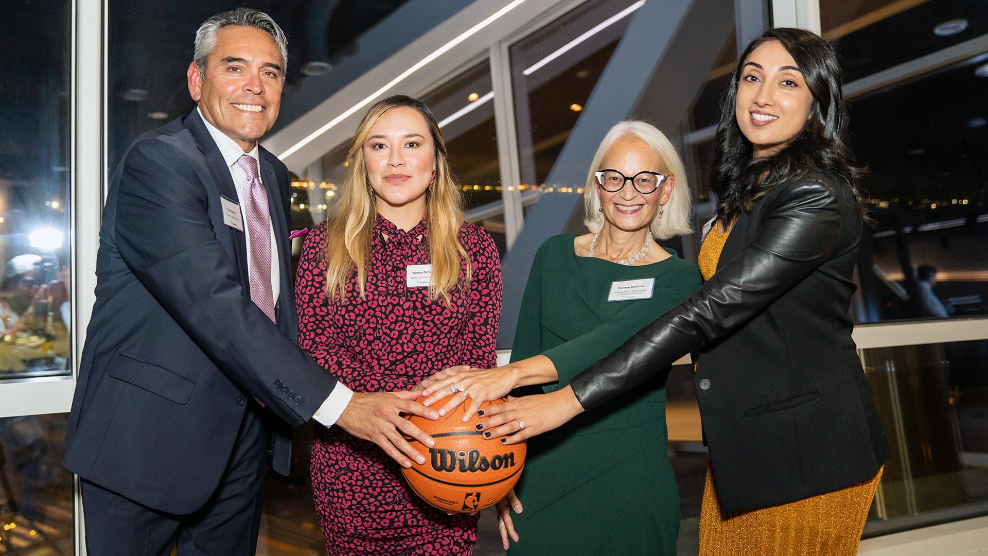 Four people each place a hand on a Wilson basketball and smile for a group photo.