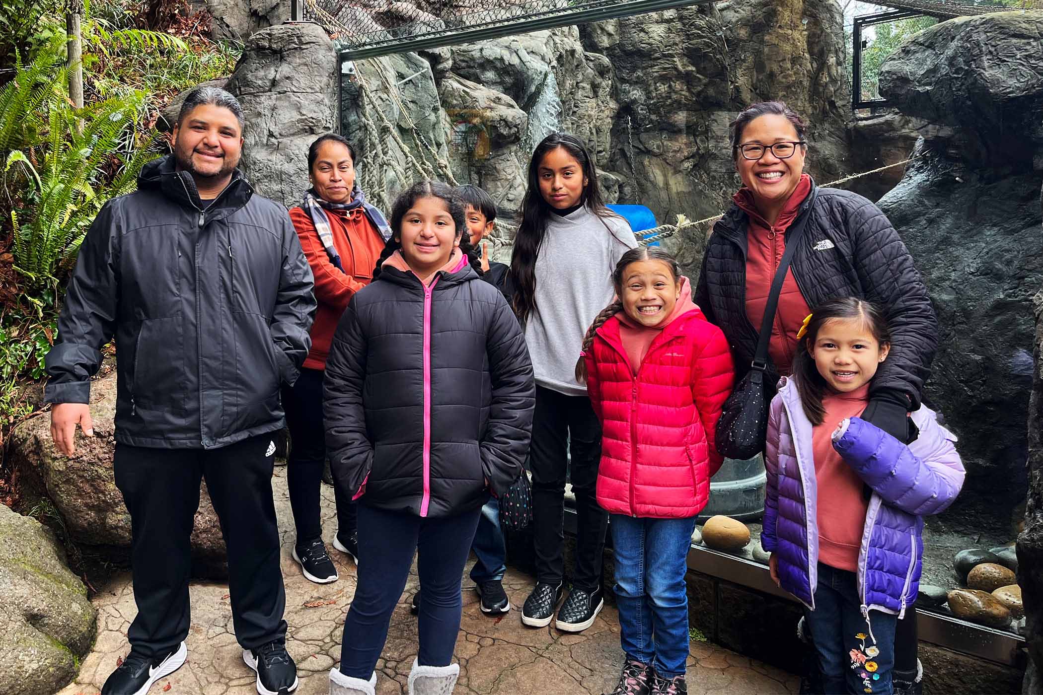 A group of eight individuals on a hike smile at camera. There are three adults and five children. In the background are rocks and vegetation.