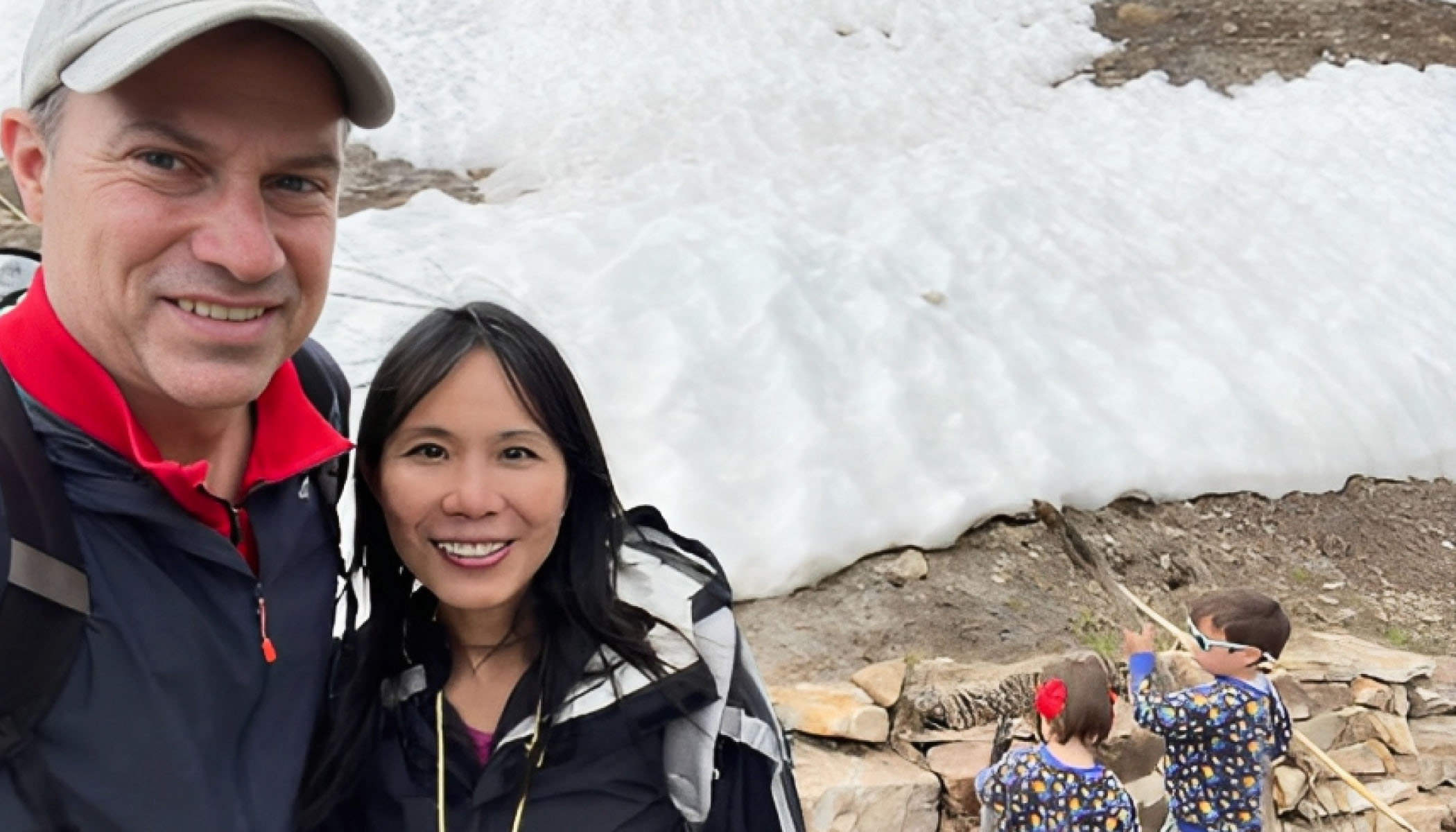 Vivian Wu standing next to her husband, both wearing jackets in front of an icy hill with their two small children playing in the background.