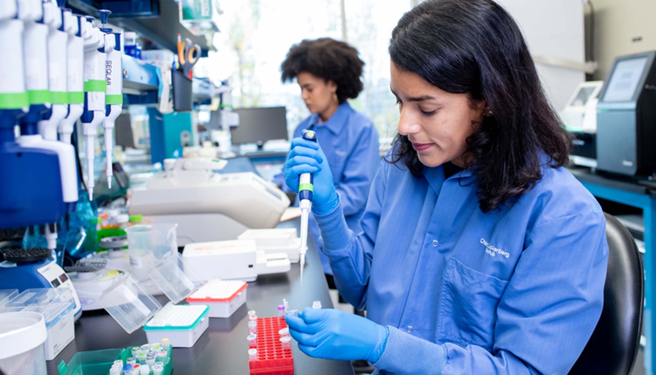 Two scientists in a lab — one scientist is holding a pipette in one hand and a test tube in the other hand.