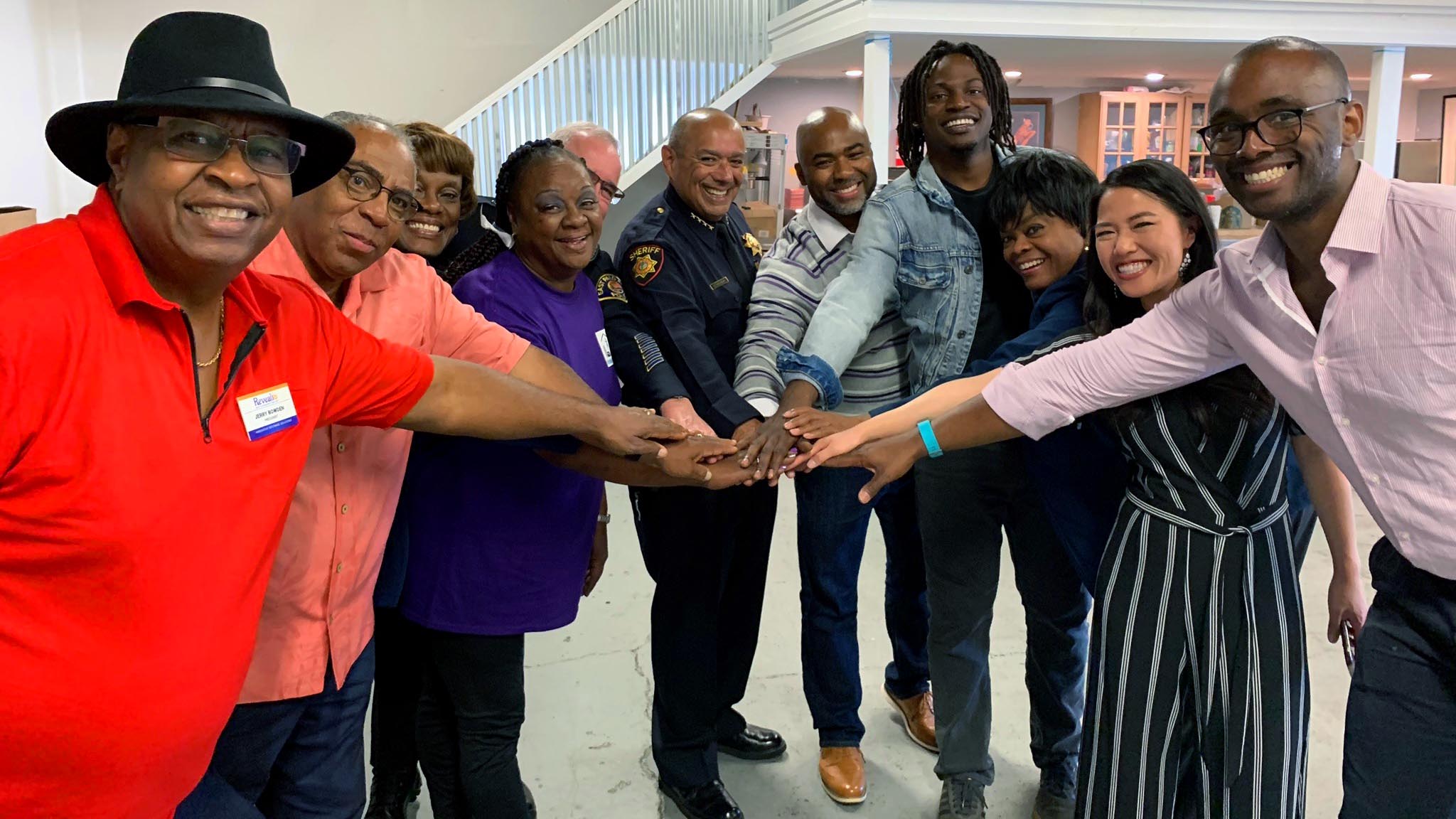 A group of smiling adults stand in a half circle with their arms stretched out and hands stacked together.