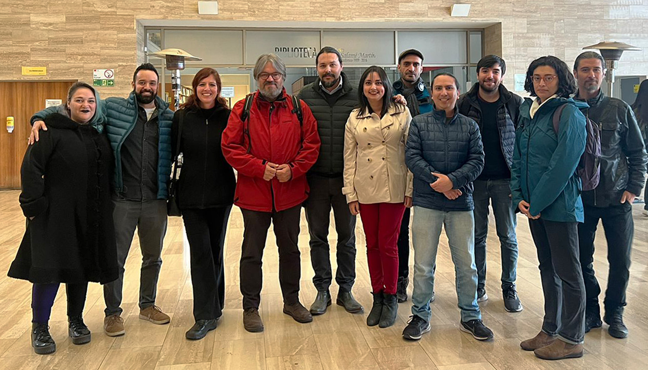 A group of people in outerwear pose for a picture. They are inside, and a sign reading “biblioteca” or library, is in the background.