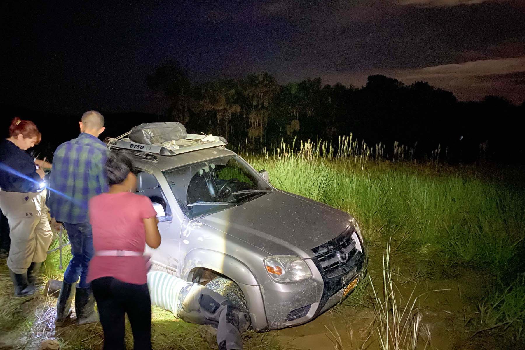 Blurry figures stand in the dark, shining a light on a gray truck. Two people kneel to fix a flat tire.