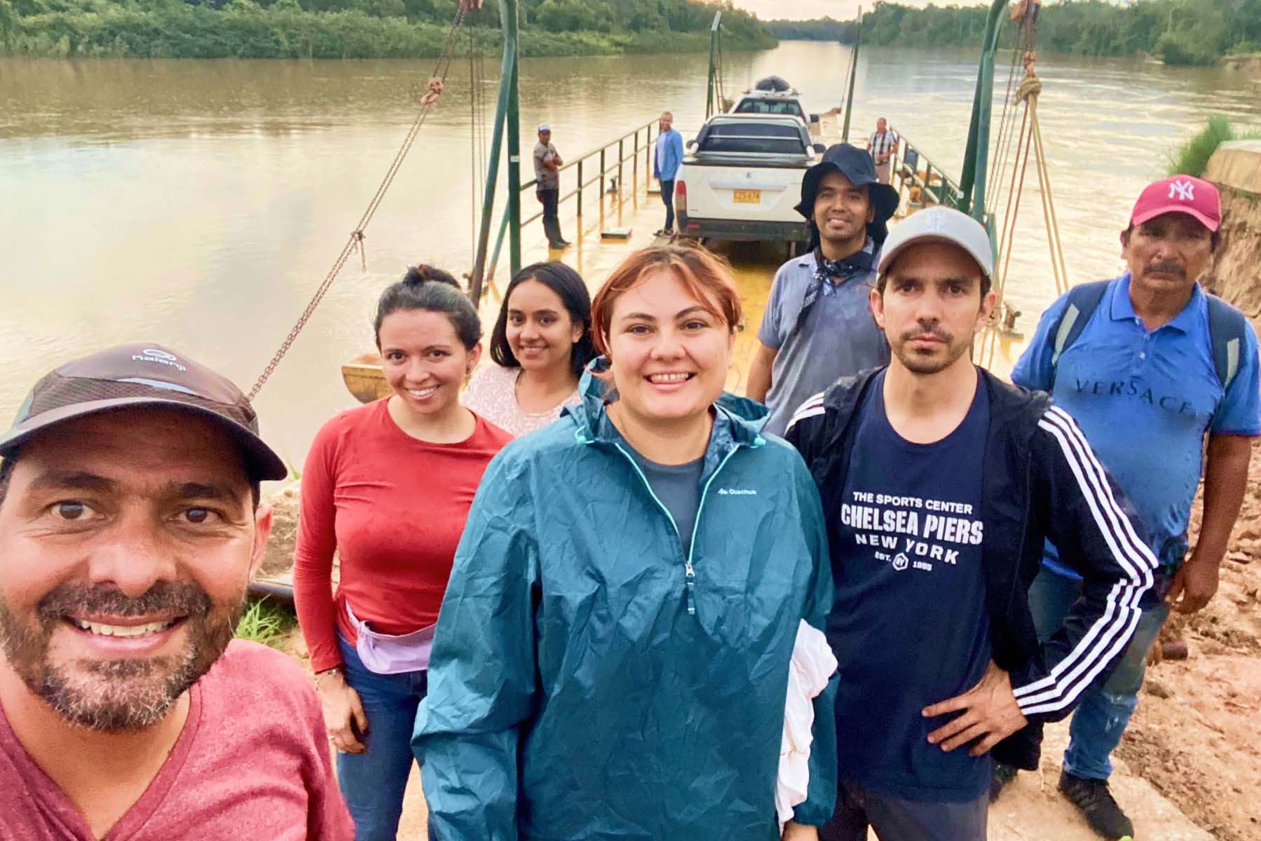 A group of people pose for a picture on a river bank. Vehicles are parked on the ferry behind them.