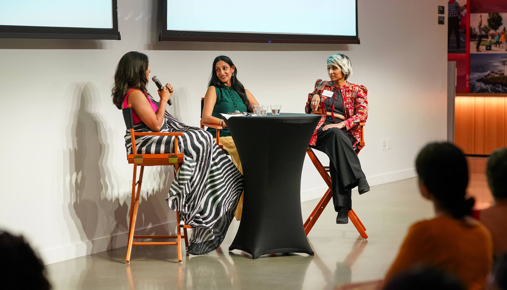 A group of women, one holding a microphone, chat at a small black table.