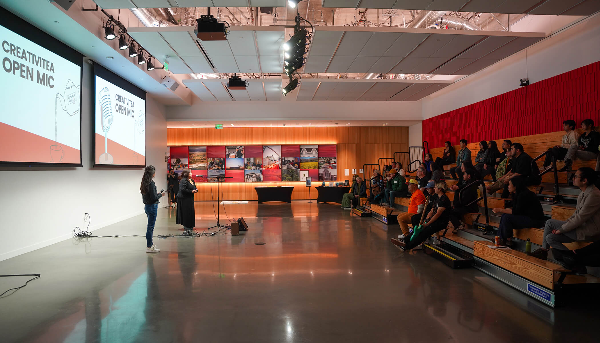 People sit on bleachers in the CZI Community Space listening to two people talk. Screens that say “CreativiTea Open Mic” are behind the speakers.