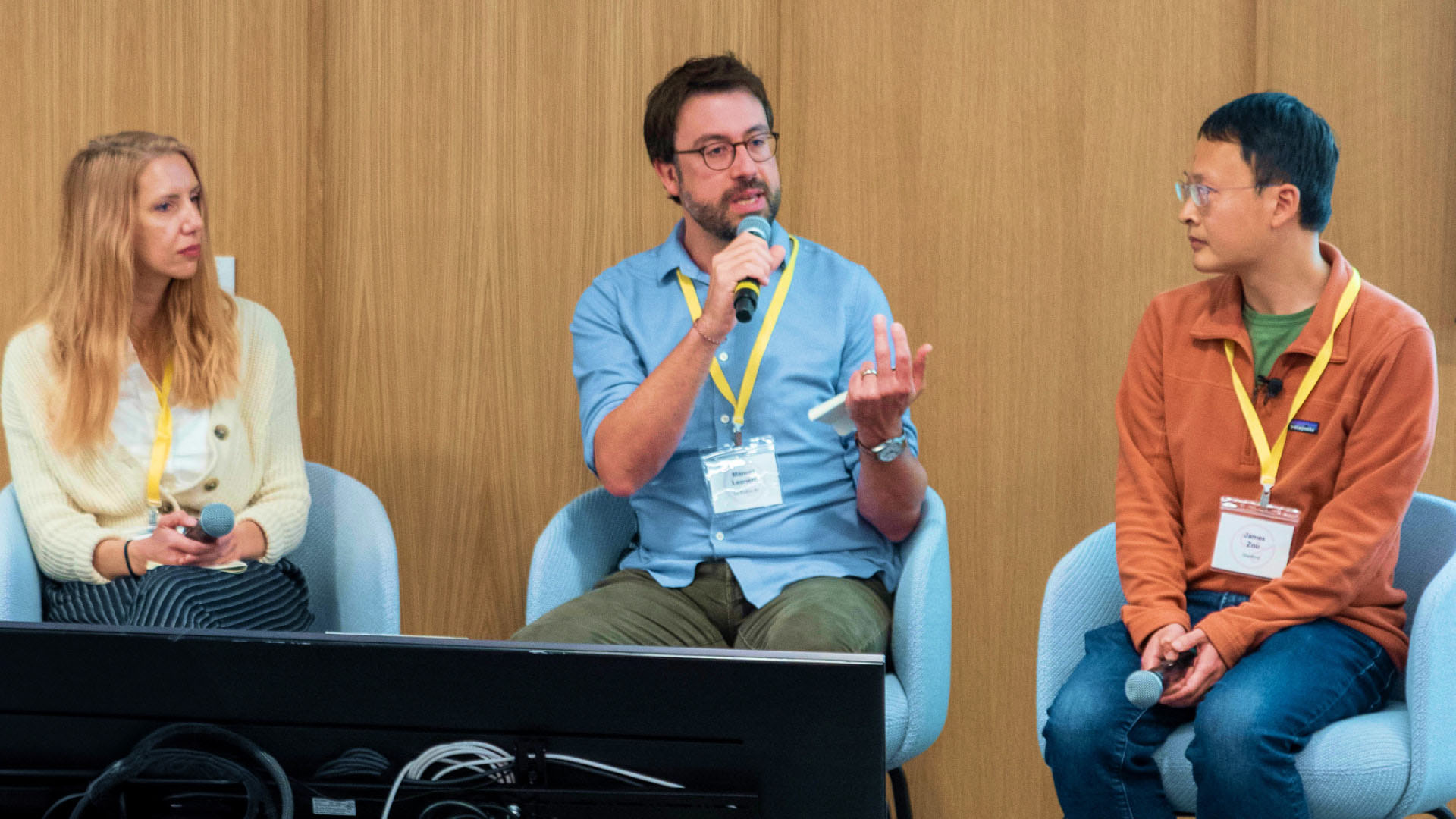Three people are seated on a panel, engaged in a discussion; a man in the middle speaks into a microphone while gesturing, with a woman on the left and a man on the right listening attentively.