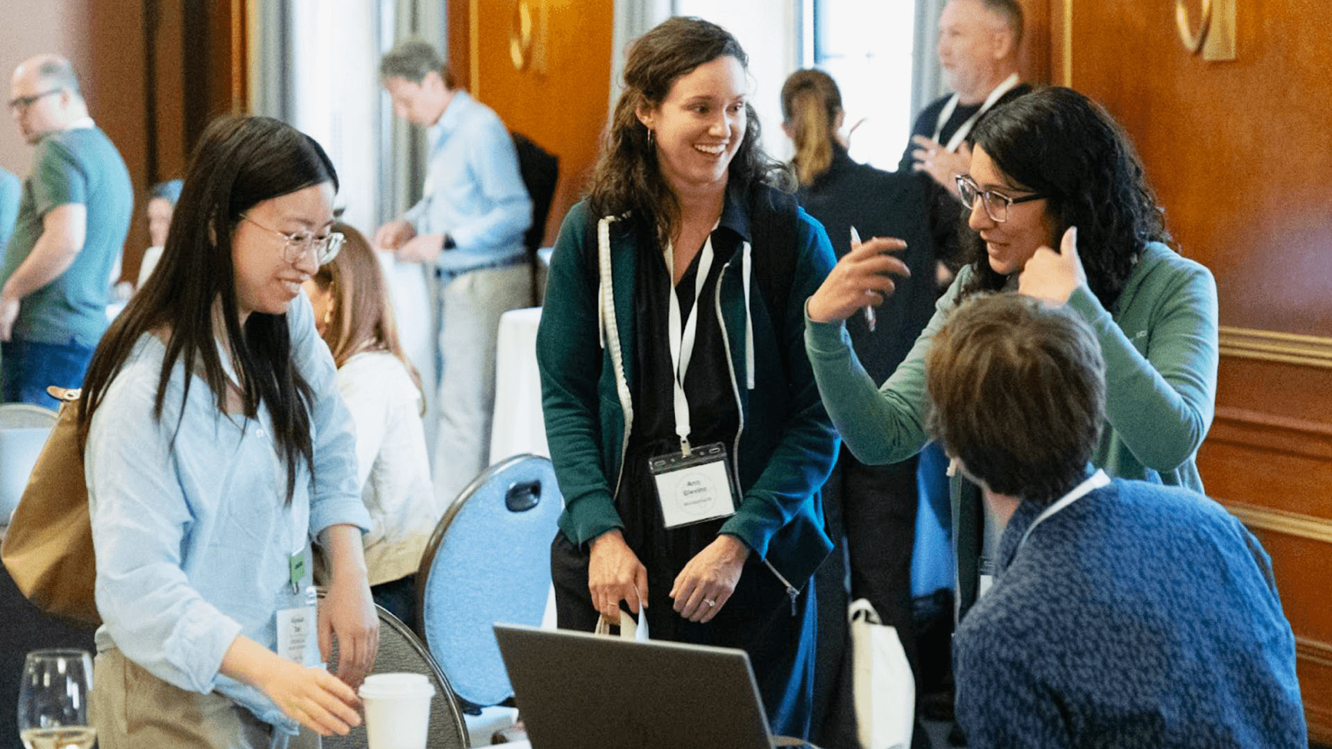 A group of people gathered around a table with a laptop, engaging in a lively discussion and smiling, at a conference.