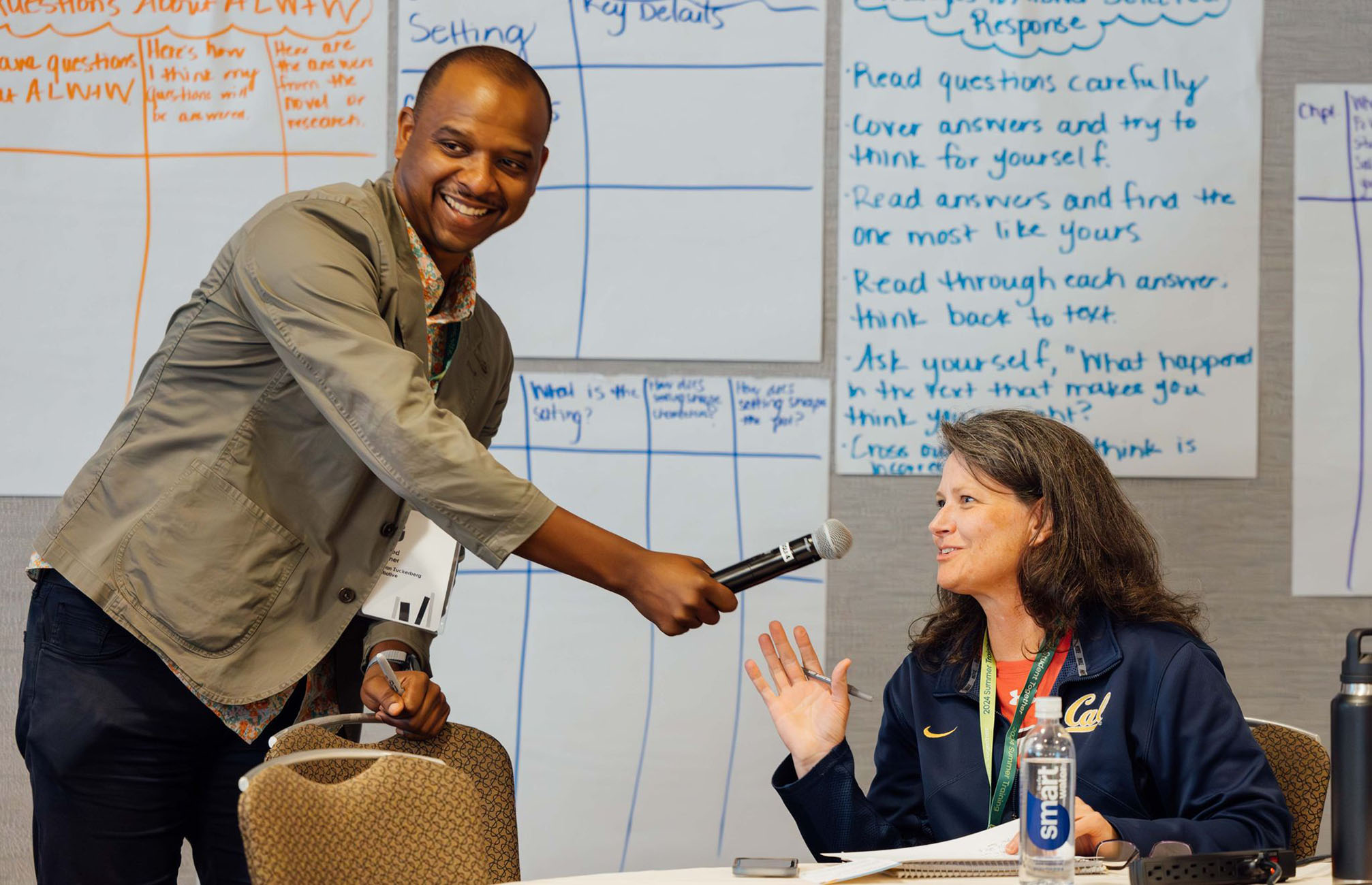 A man standing holding a microphone for a woman sitting at a desk