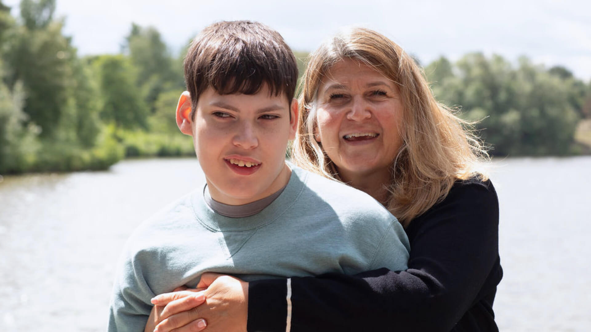 A smiling woman stands behind her son. They are outdoors near a body of water.