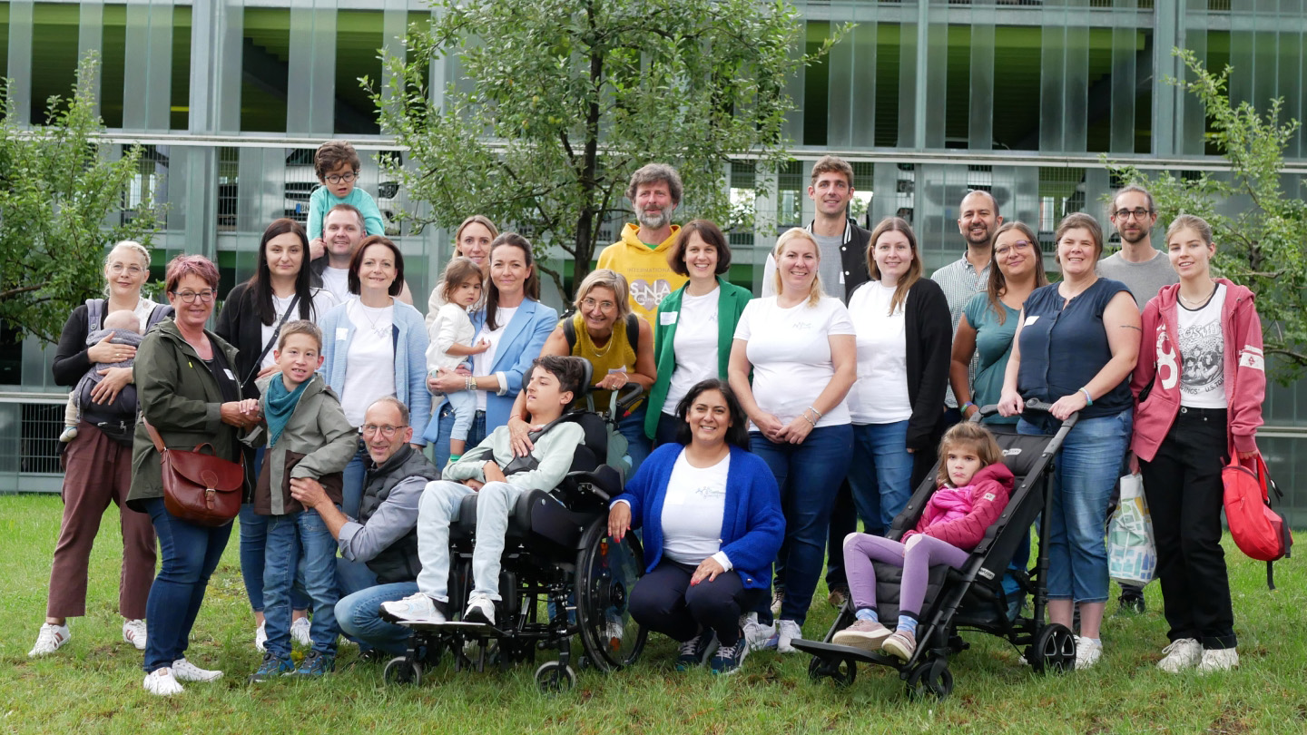 A diverse group of adults and children, including individuals in wheelchairs and strollers, gather together outdoors in a grassy area for a group photo. The group smiles warmly at the camera, and the background features trees and a modern building with glass paneling.