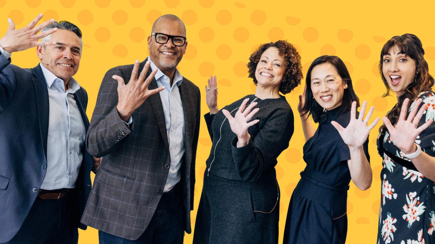 A group of five individuals smiling and holding up five fingers in front of a bright yellow background with a dotted pattern.